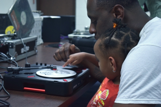 A black man plays with a turntable, with his daughter in his lap